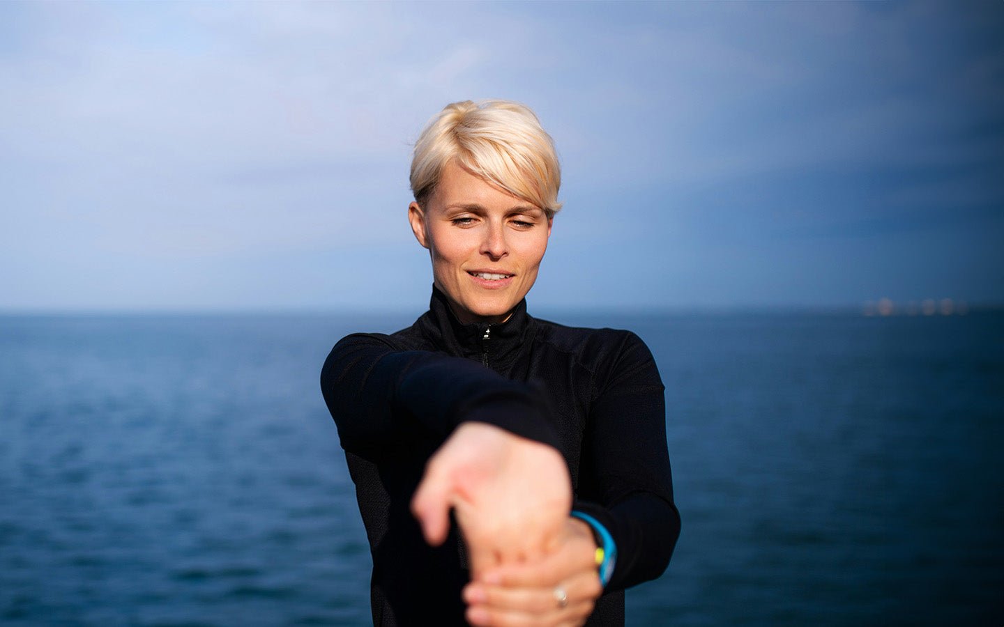 Blonde woman in black athletic top stretching arms by the ocean