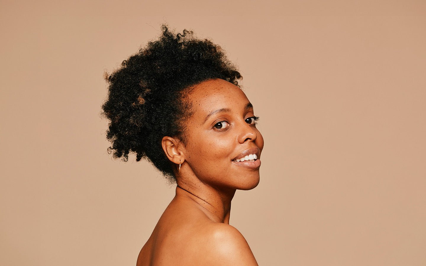 Smiling woman with dark curly hair and bare shoulders against a beige background