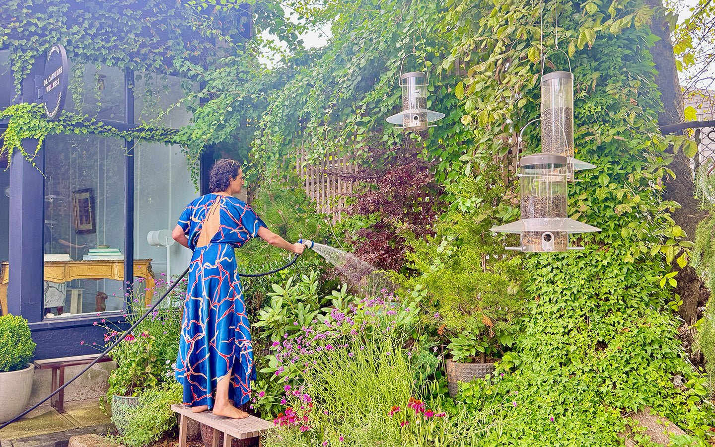 Woman in blue dress watering lush garden with bird feeders