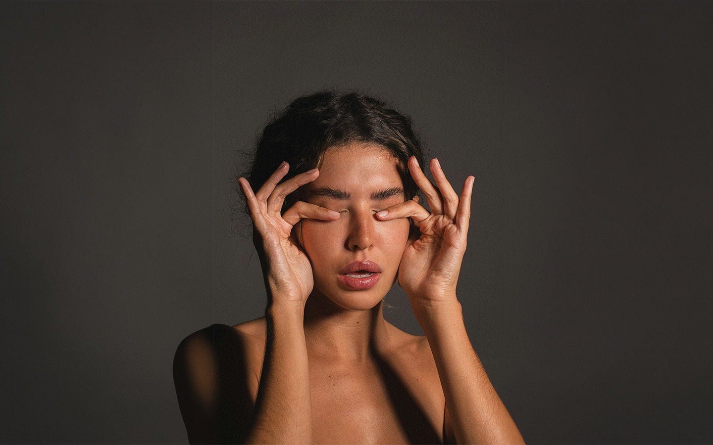 Woman with closed eyes, fingers gently pressing on eyelids, against a dark gray background