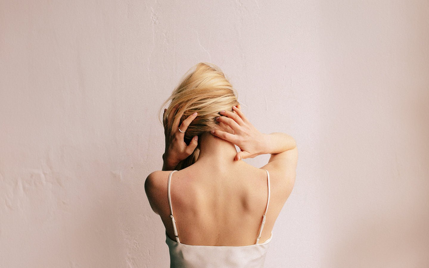 Blonde woman from behind, hands on neck, wearing a white camisole against a light wall