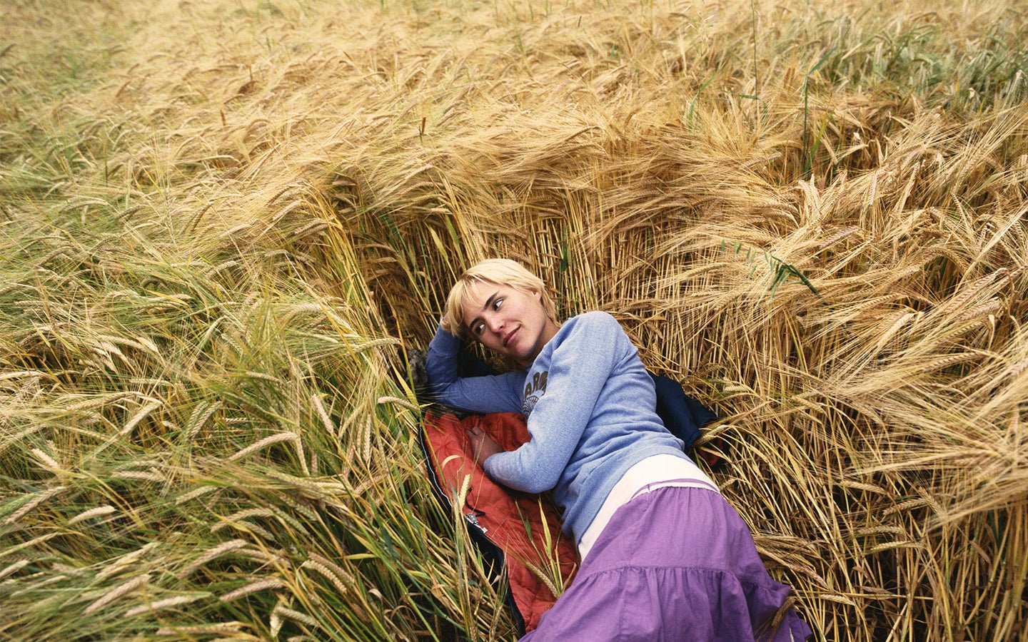 Blonde woman in blue top and purple skirt lying in a golden wheat field