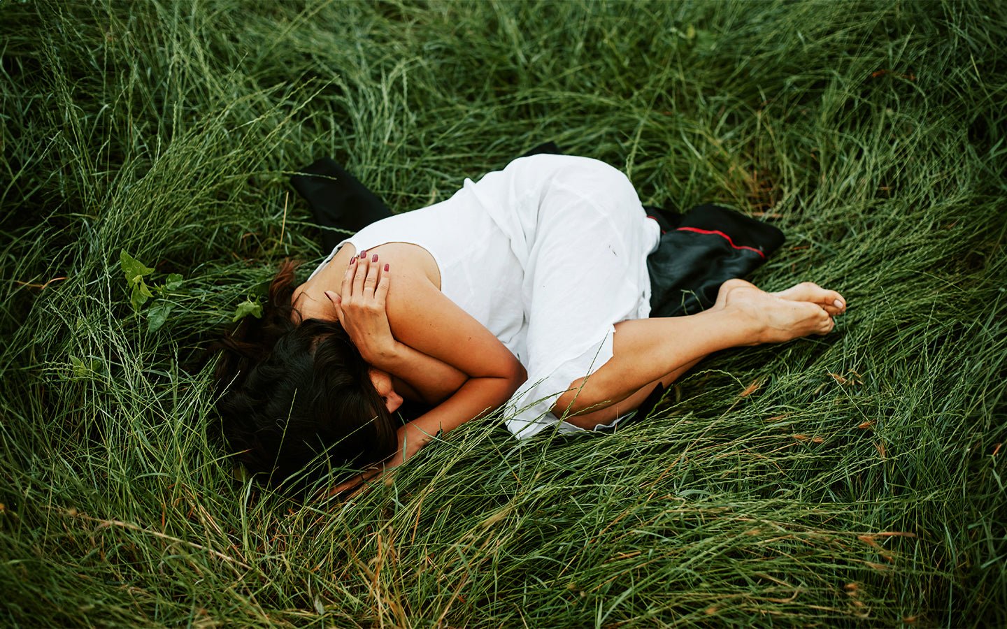 Woman in white dress curled up on green grass, hands covering face