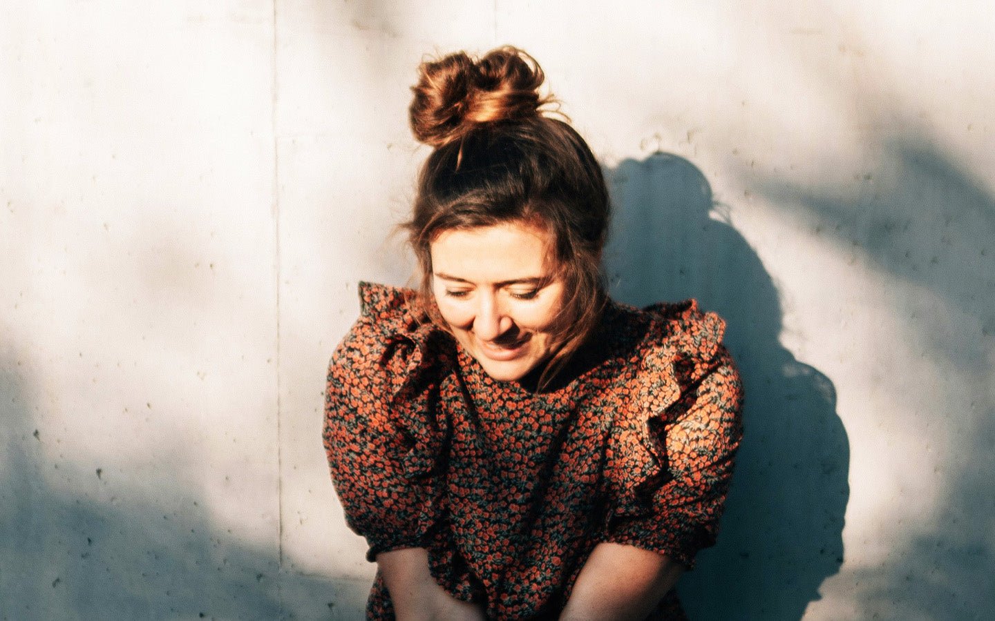 Woman with top bun, floral dress, smiling, against a white wall with shadows