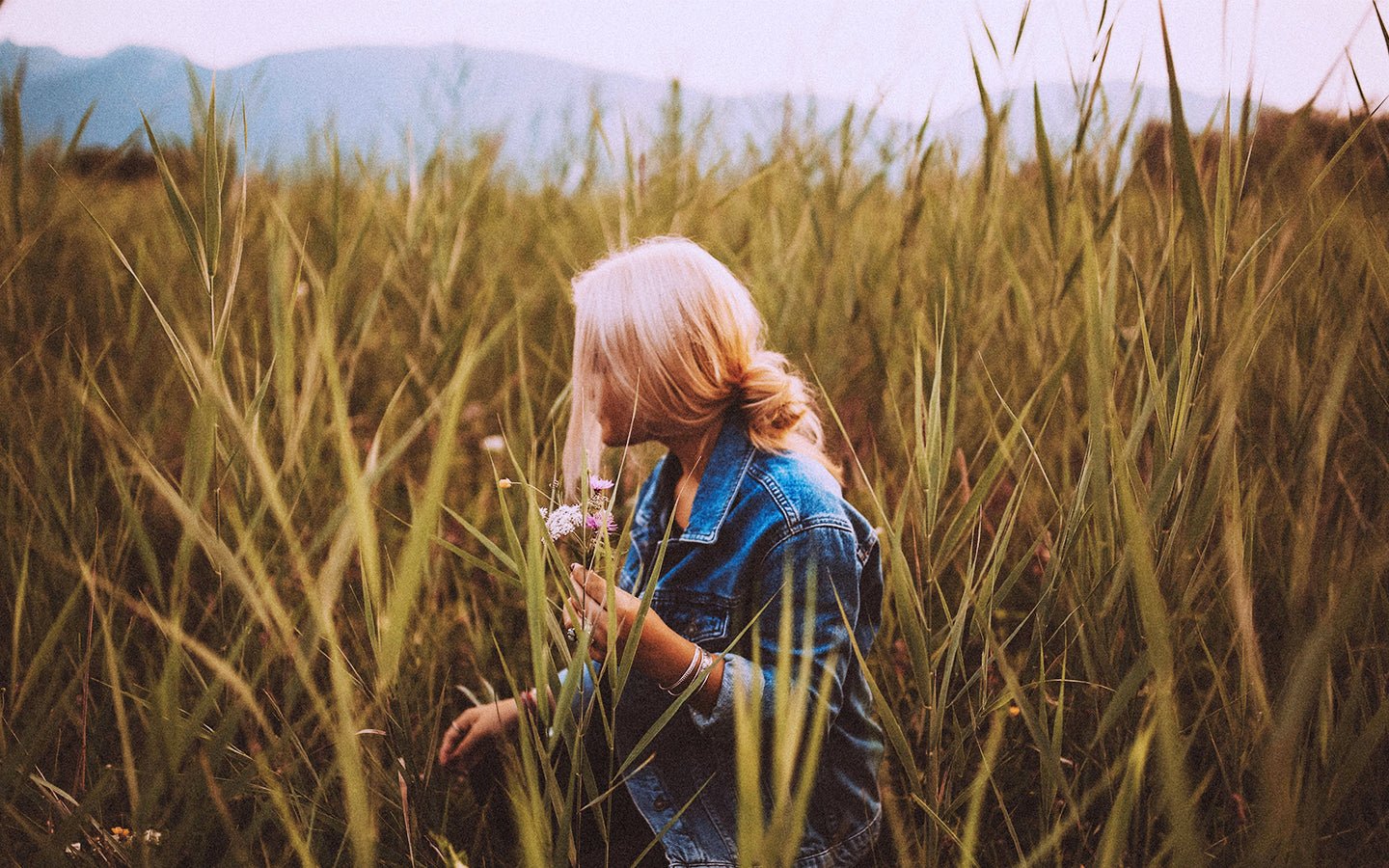Blonde woman in denim jacket picking flowers in a field of tall grass