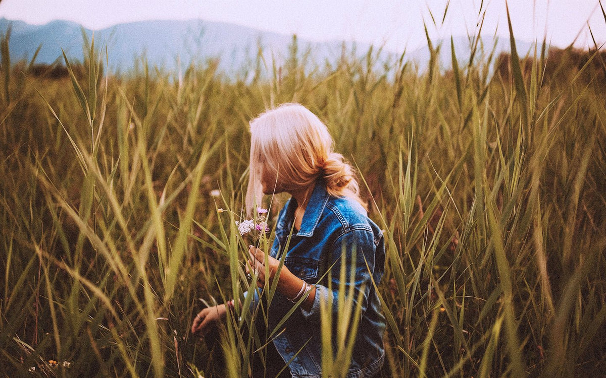 Blonde woman in denim jacket picking flowers in a field of tall grass
