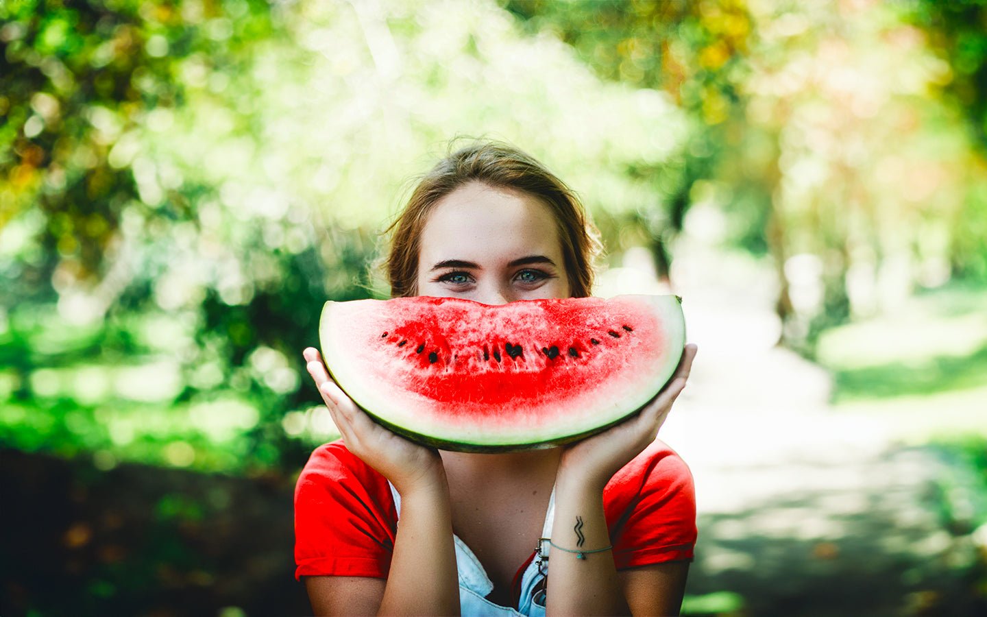 Woman in red shirt holding a slice of watermelon, smiling in a green park