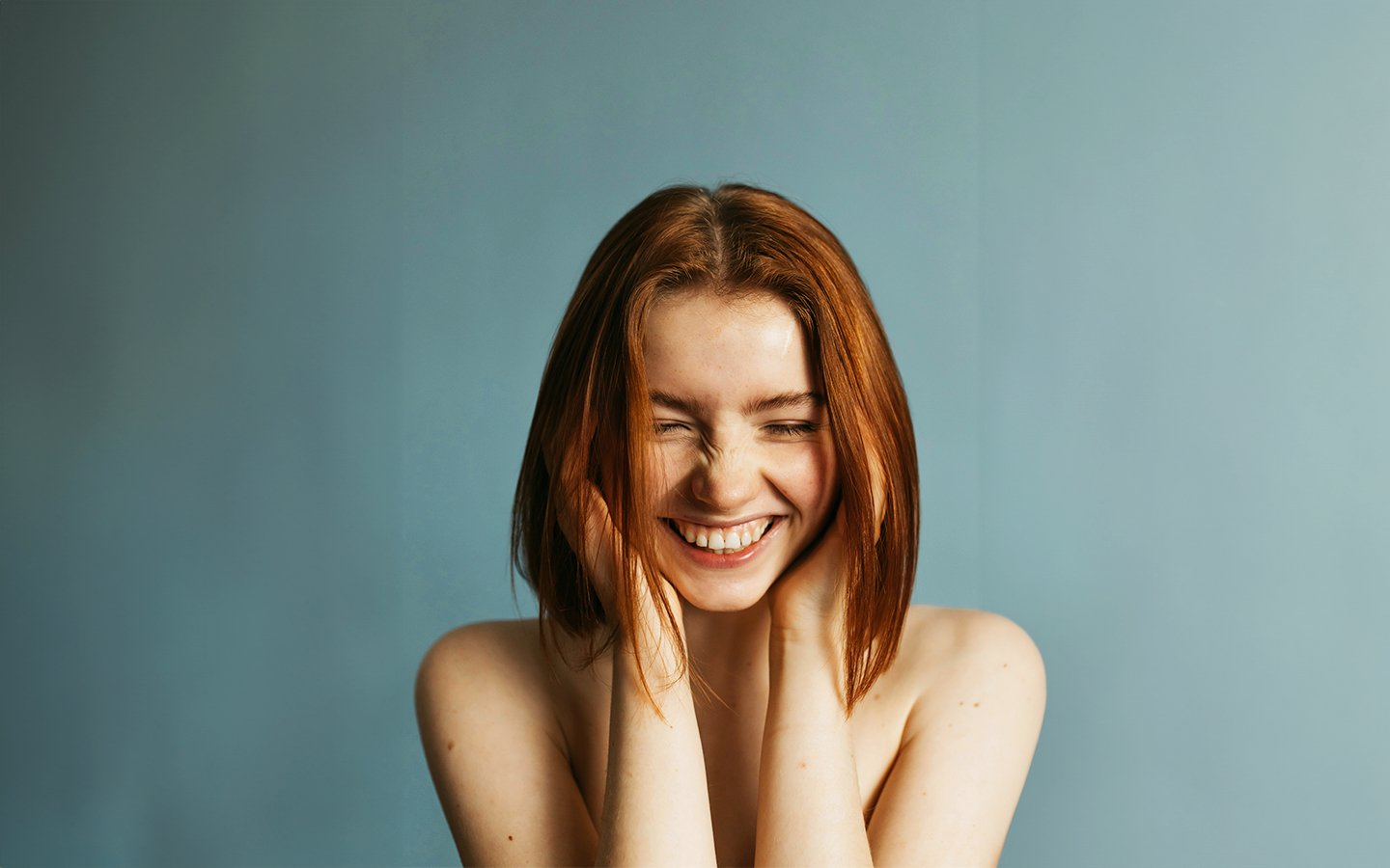 Smiling woman with red hair, bare shoulders, and hands framing her face against a blue wall