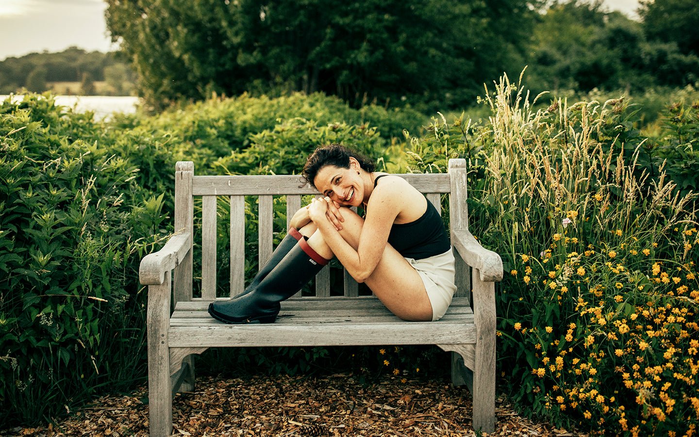 Woman in black tank top and boots smiles on a park bench surrounded by greenery