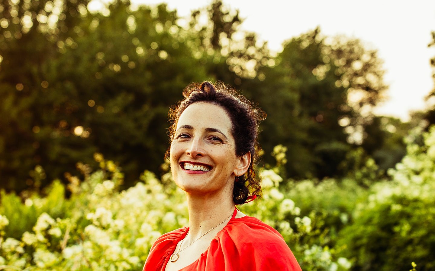 Smiling woman in red top with curly hair in a sunny green garden
