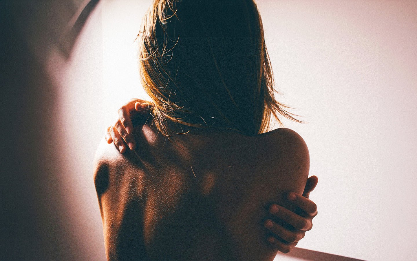 Woman from behind, arms crossed, embracing shoulders, against a light background