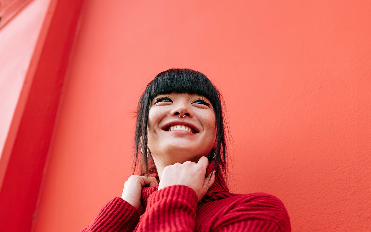 Smiling woman in red sweater with bangs and septum piercing against a coral wall
