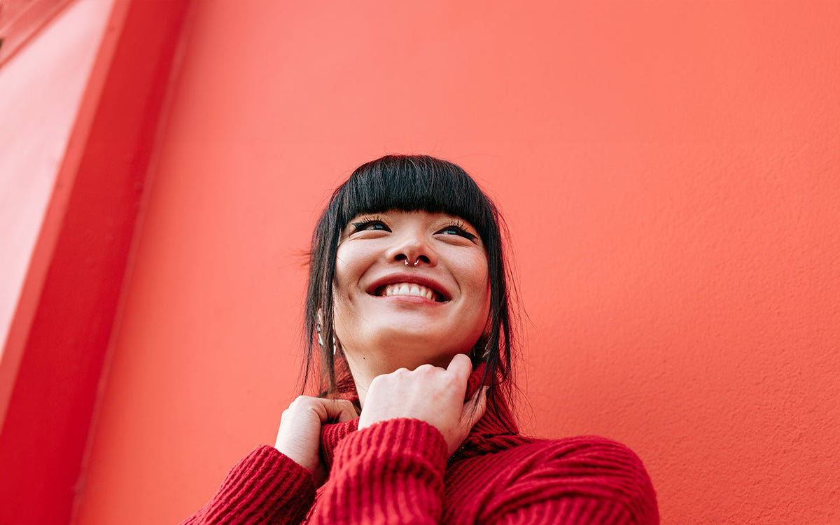 Smiling woman in red sweater with bangs and septum piercing against a coral wall