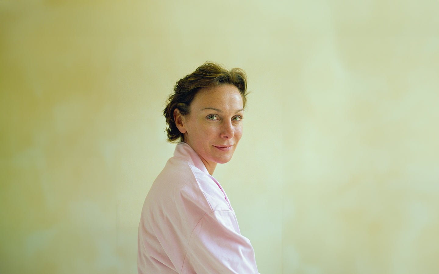 Woman with short brown hair and green eyes, wearing a pink robe, looking over her shoulder
