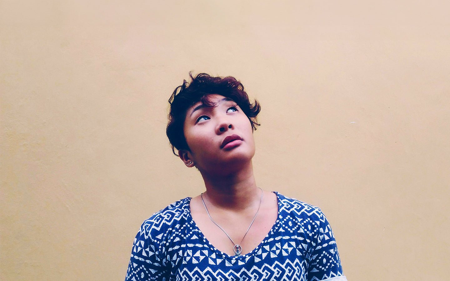 Woman with short dark hair and blue patterned shirt looking up against a beige wall