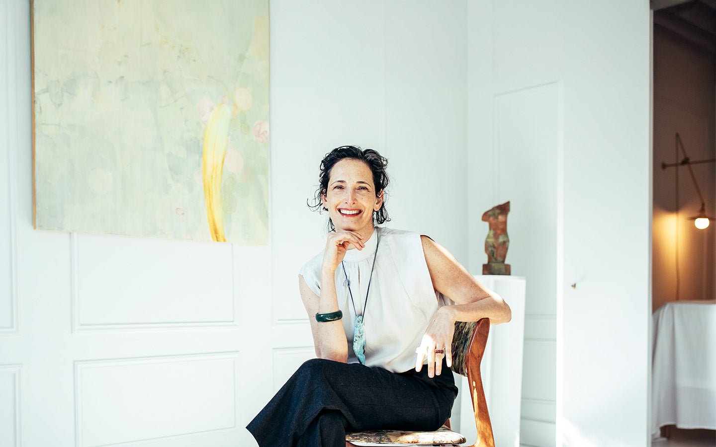 Woman with dark hair, white top, and black pants smiling in a bright room with art