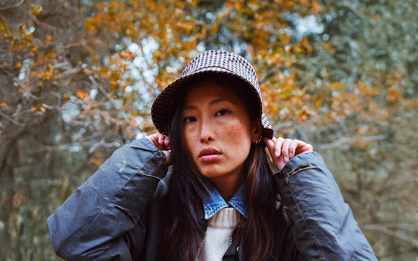 Woman in a plaid bucket hat and green jacket, hands near ears, with autumn leaves behind