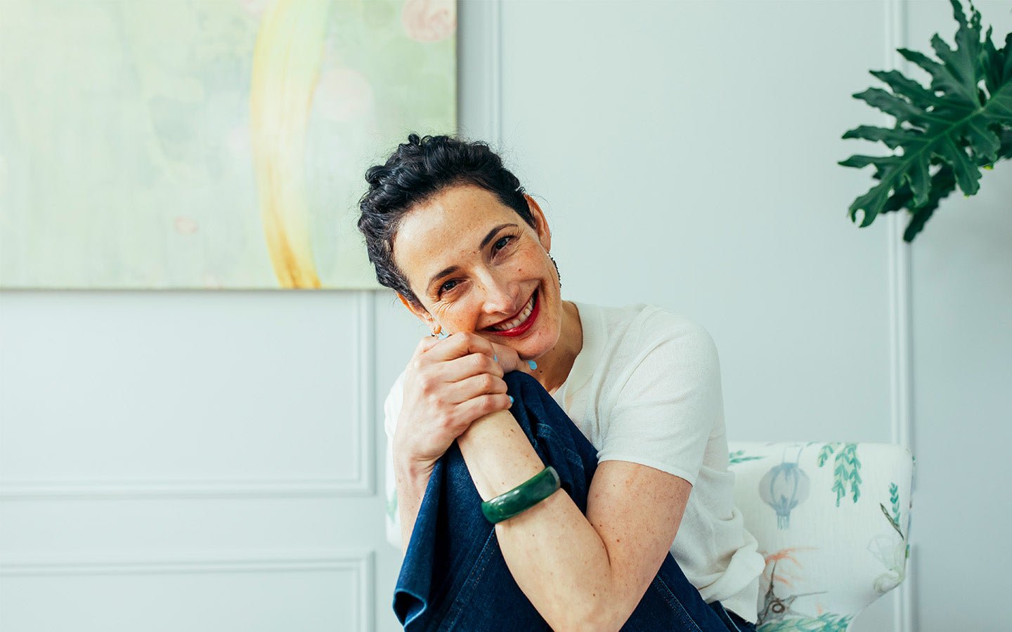 Smiling woman with dark hair, white shirt, jeans, and green bracelet, sitting indoors
