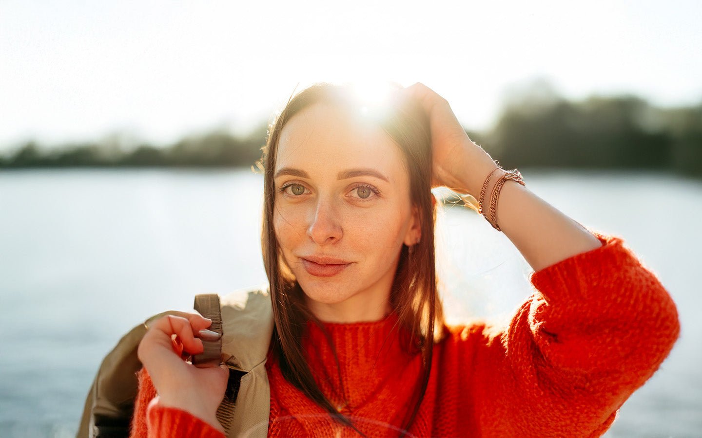 Woman in red sweater with hand in hair, sunlight on face, by a lake