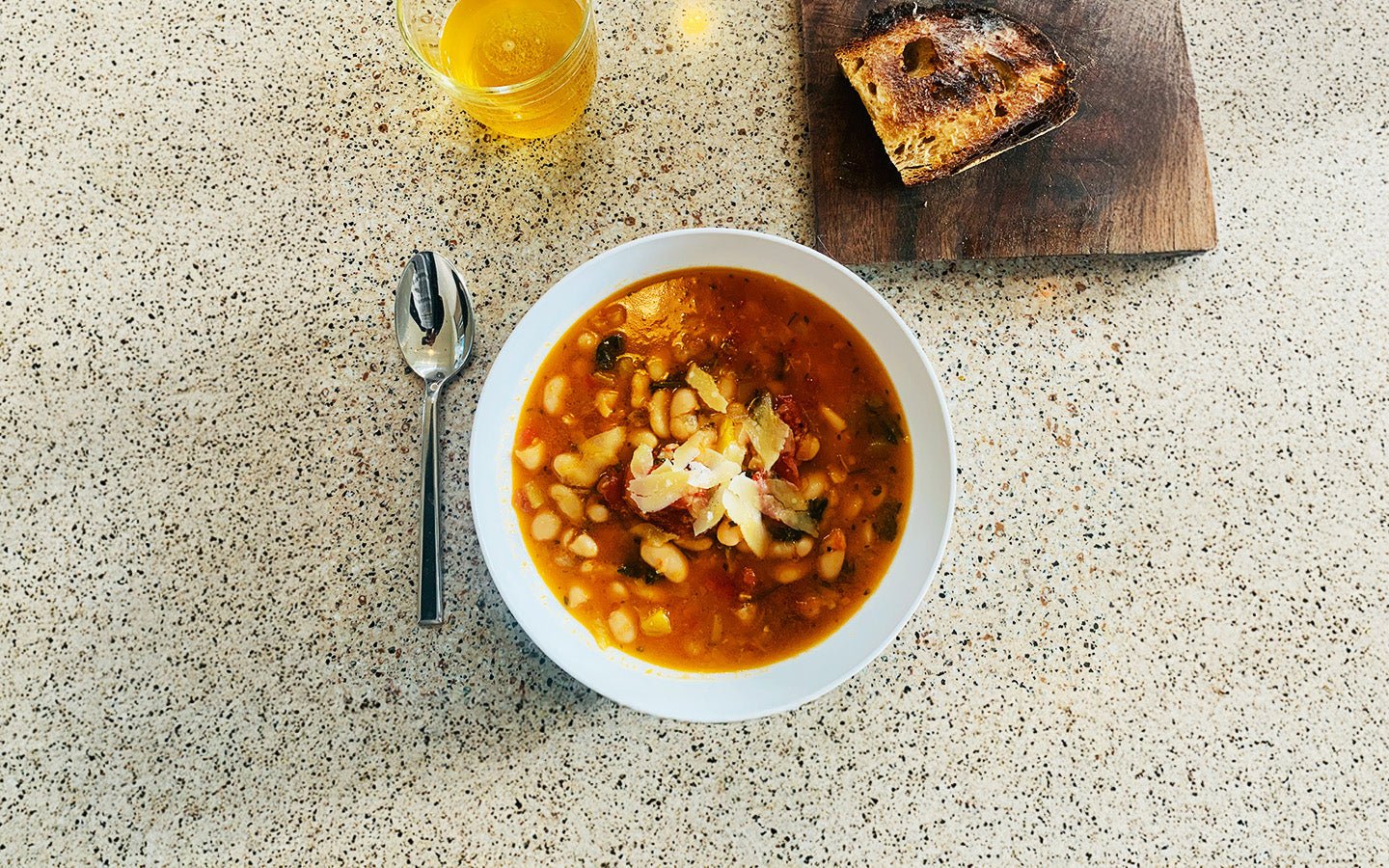 White bean soup with parmesan, spoon, drink, and toasted bread on a speckled counter