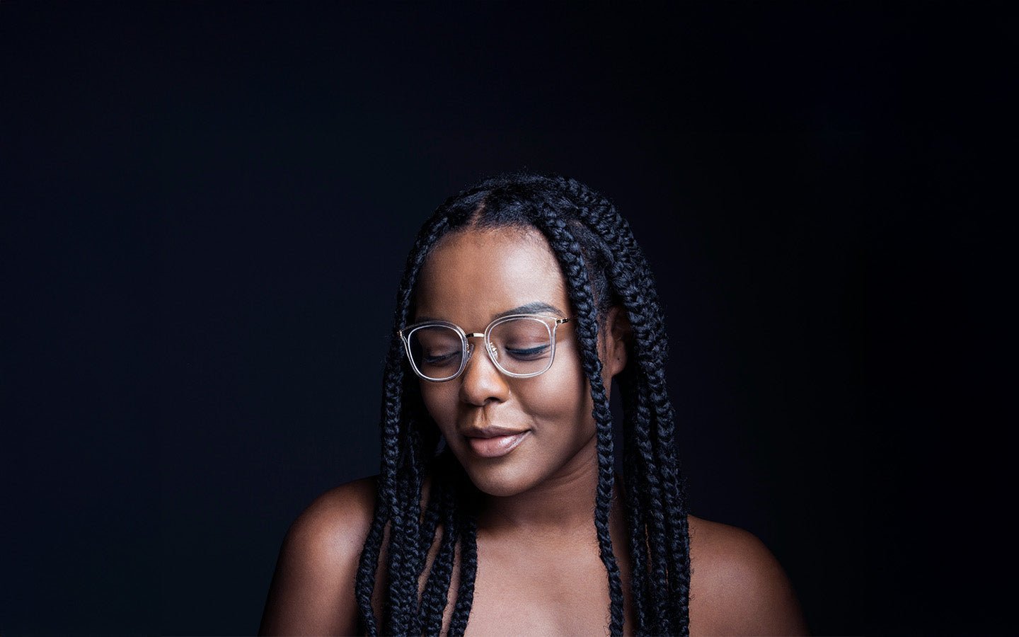 Woman with braids and glasses, eyes closed, smiling against a dark background