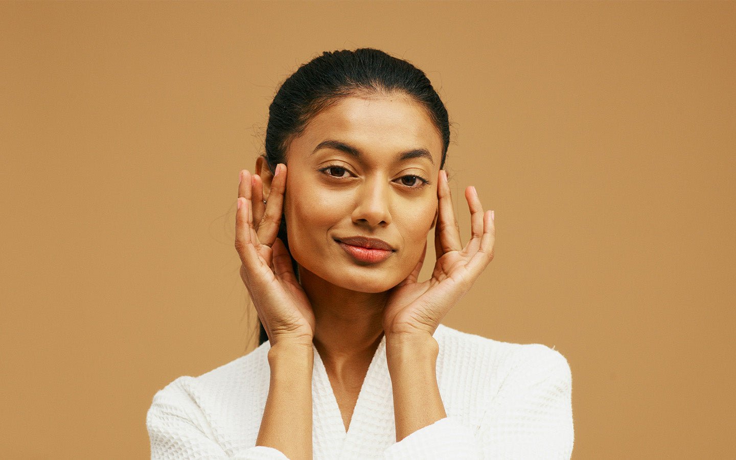 Woman in white robe with hands on face against a tan background