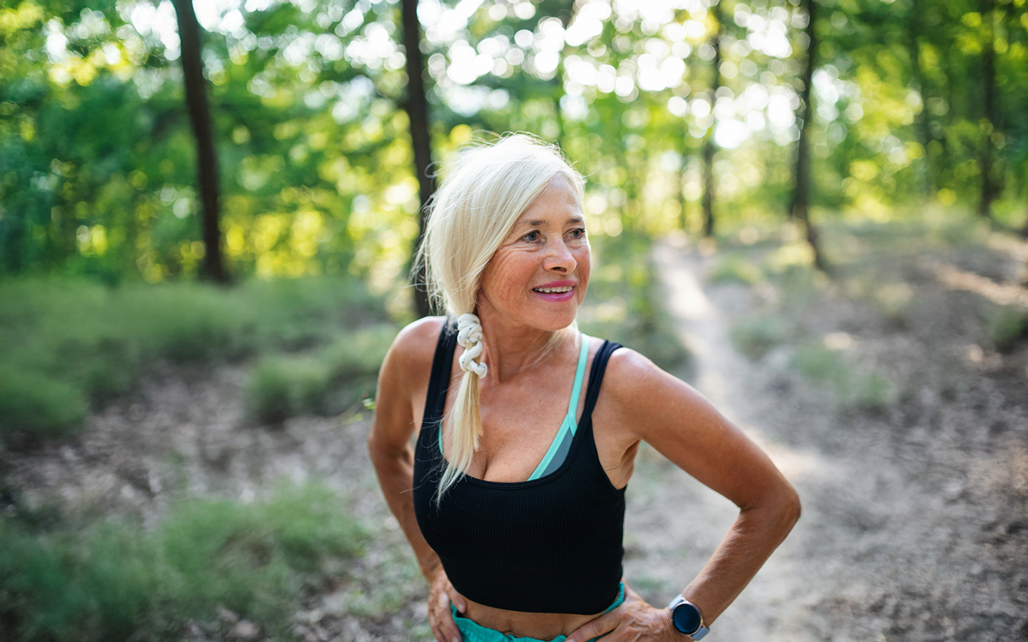 Blonde woman in black tank top and teal sports bra smiles on a forest path