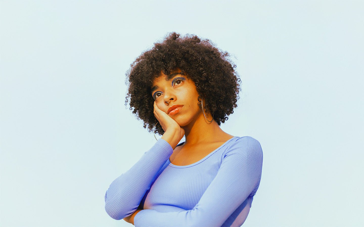 Woman with curly hair, blue eyeshadow, and nose ring, resting chin on hand