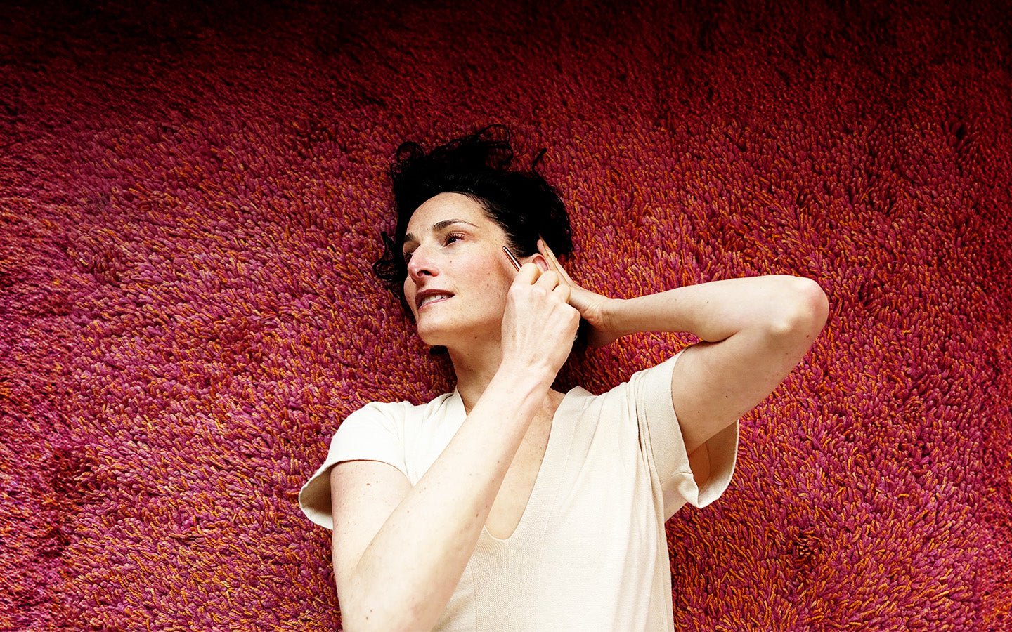 Woman in cream shirt lies on a red shag rug, using a gua sha tool on her face