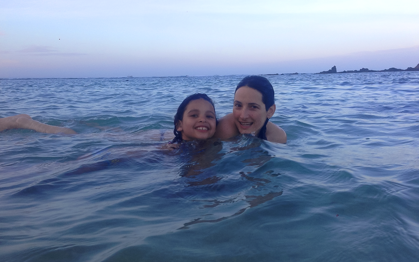 Dr. Catherine and her young daughter Ava, cheek to cheek and smiling, swimming together in the ocean at dusk in Brazil