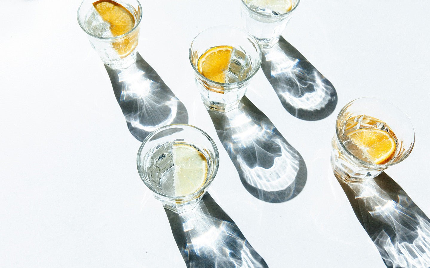 Five glasses of water with citrus slices and ice, casting long shadows