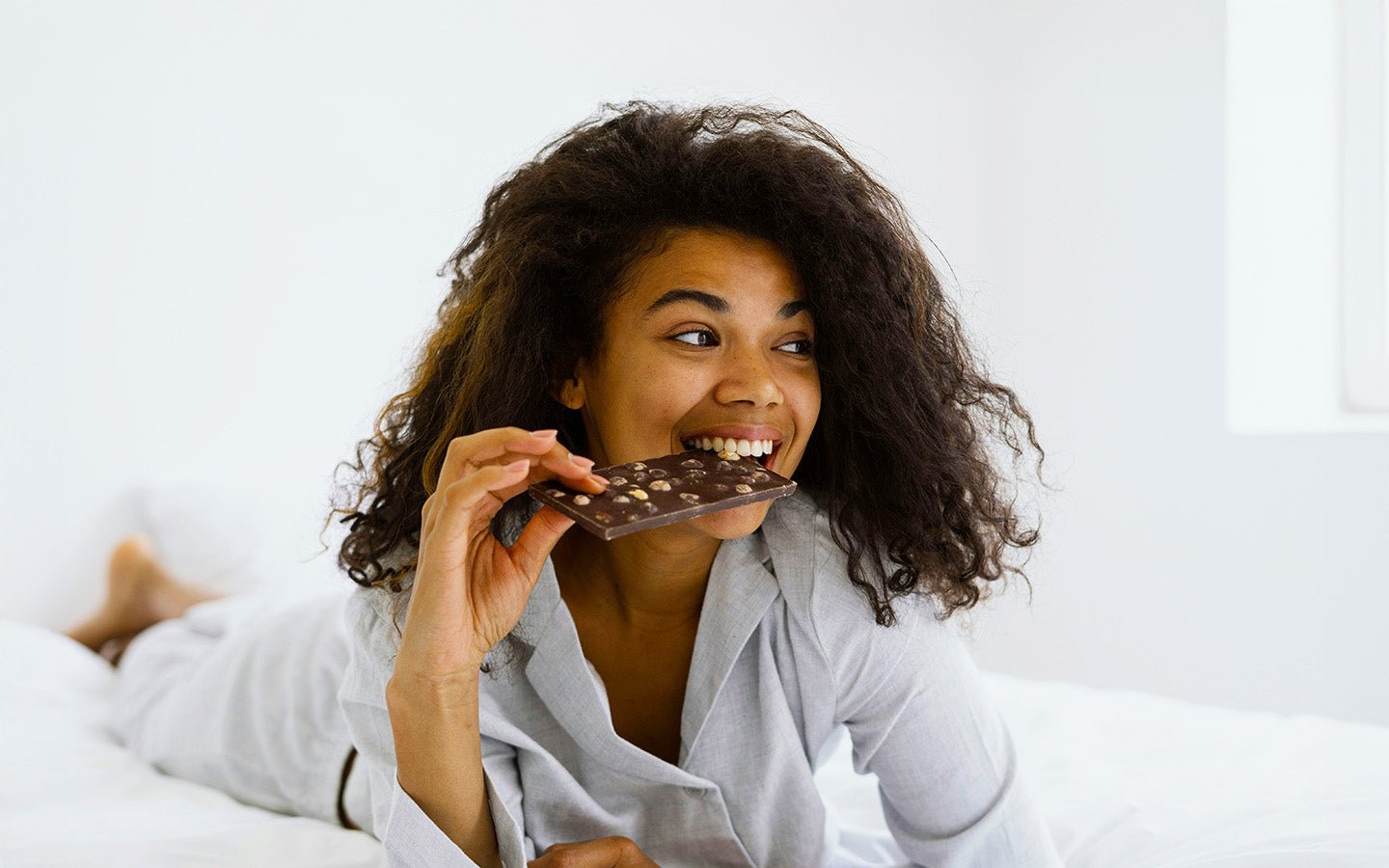 Woman with curly hair eating a chocolate bar with nuts in bed