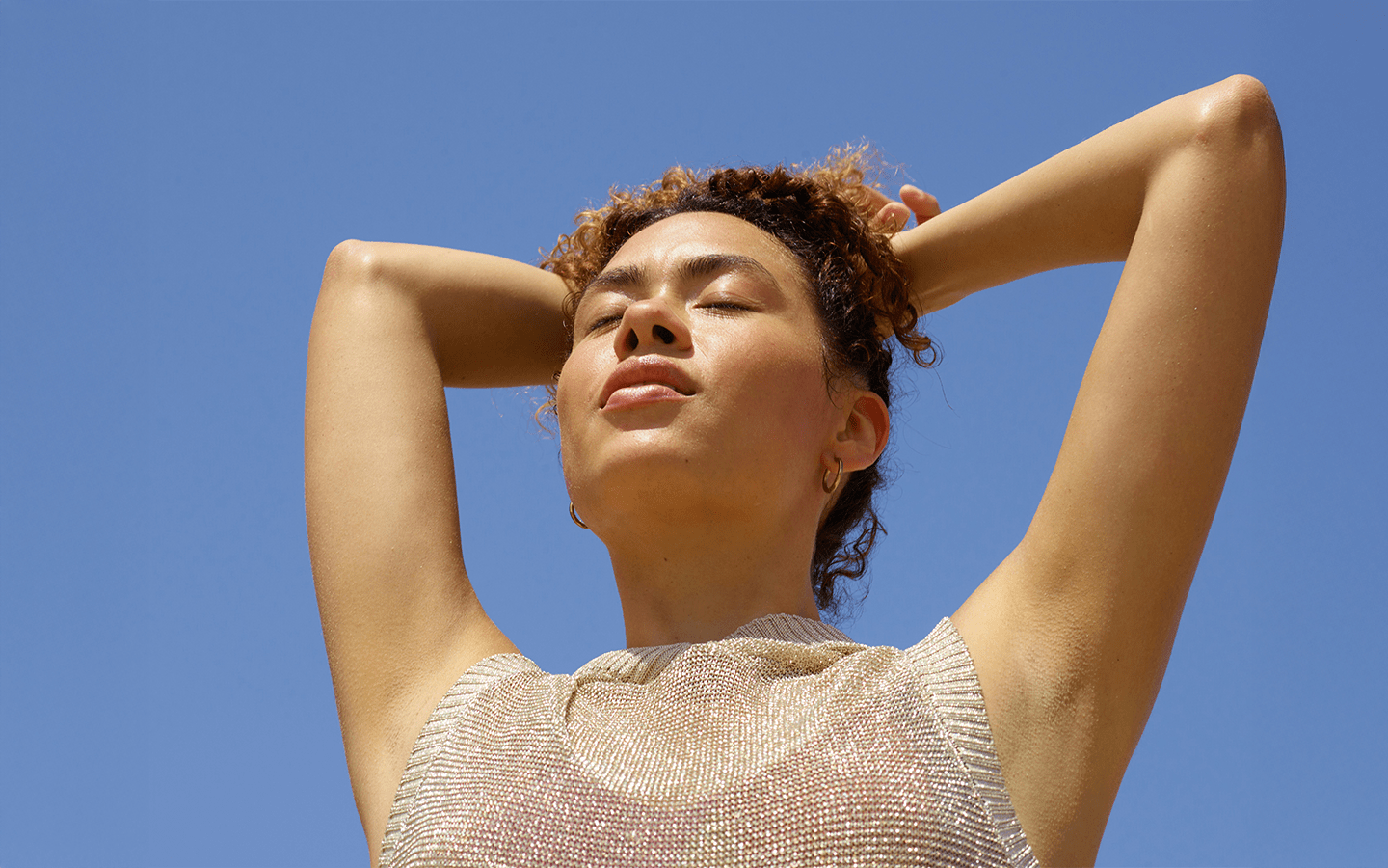 Woman with eyes closed and arms raised behind her head against a clear blue sky