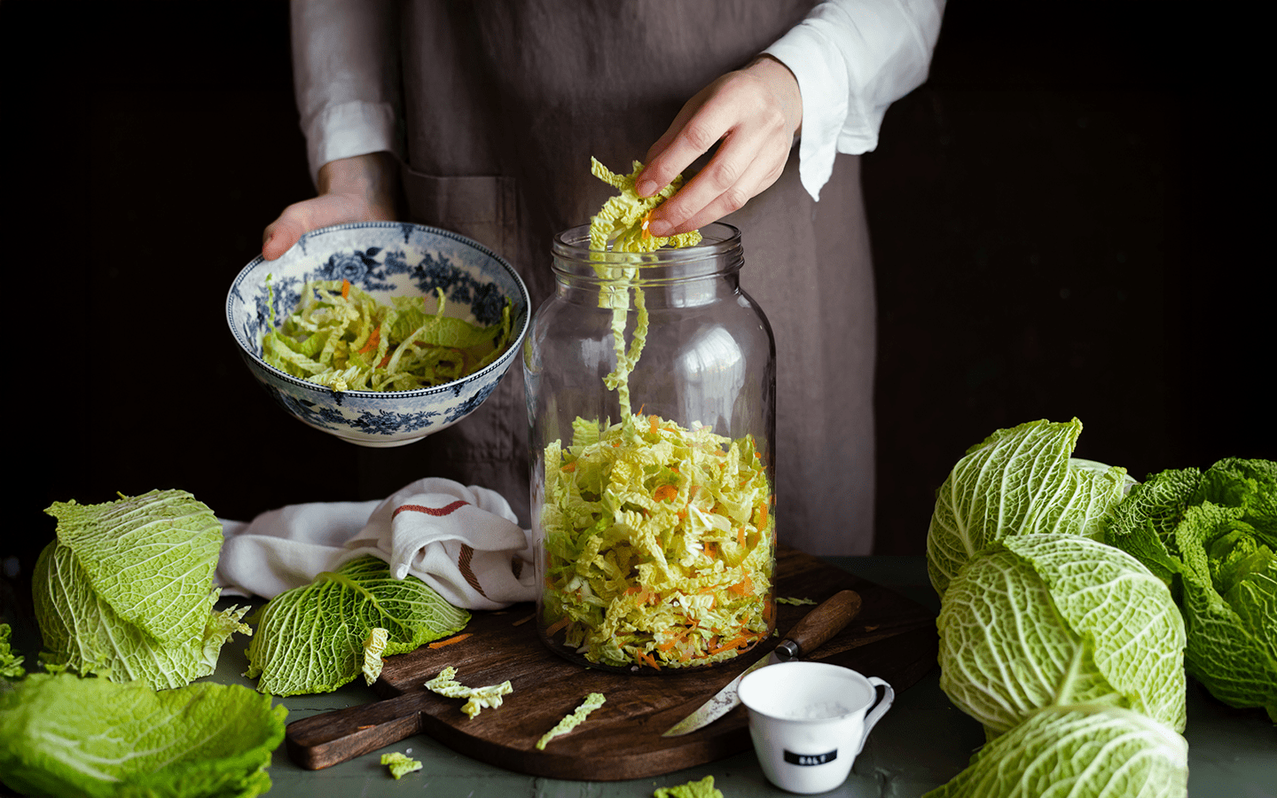 Woman in apron adding chopped cabbage to a jar, with whole cabbages and salt nearby