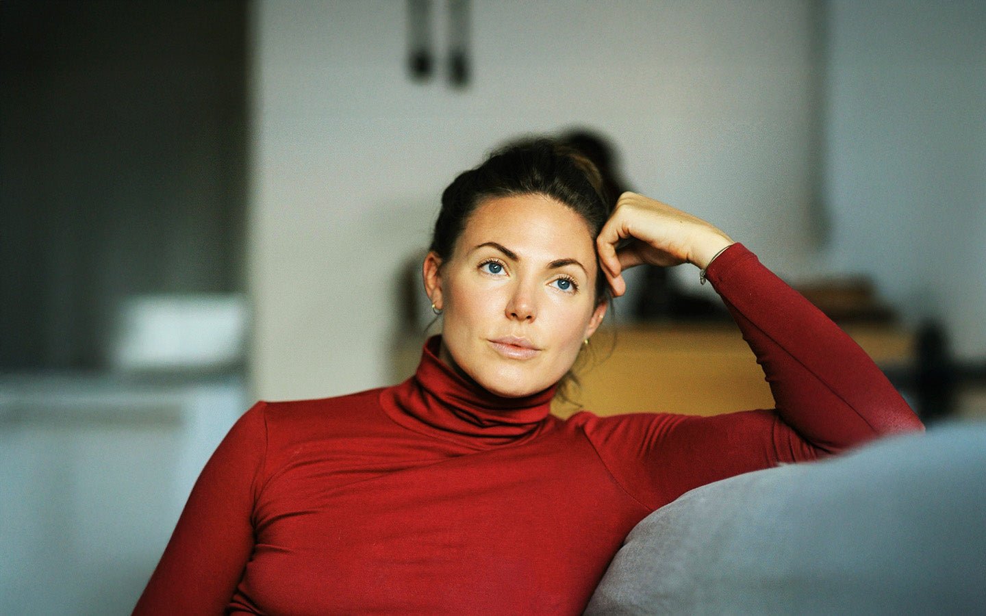 Woman in red turtleneck, blue eyes, resting head on hand, on a gray couch