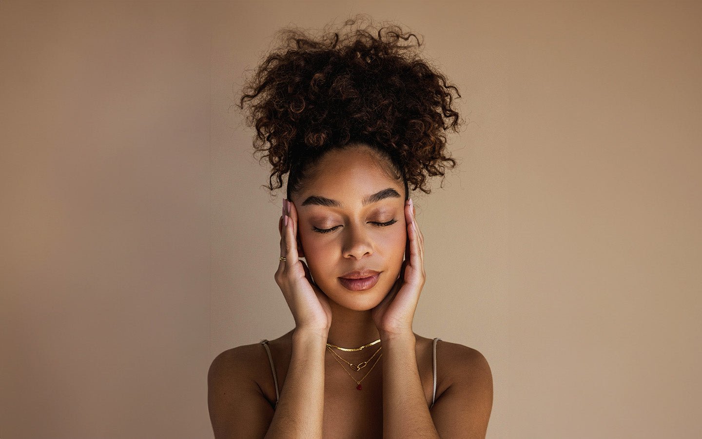 Woman with closed eyes, hands on temples, wearing gold necklaces against a beige background