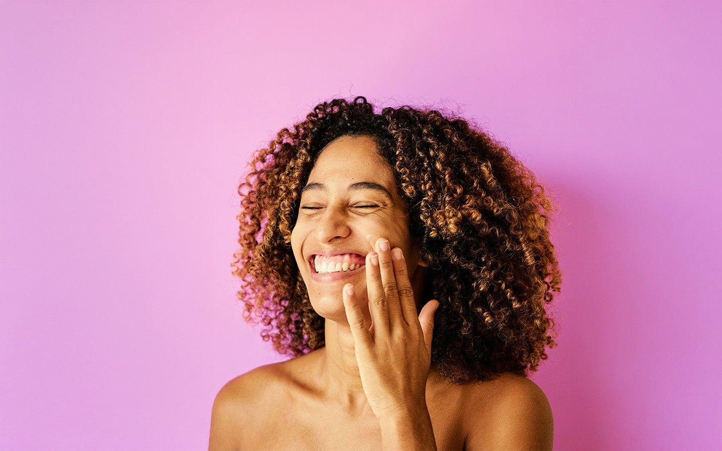 Smiling woman with curly hair applying cream to her cheek against a purple background