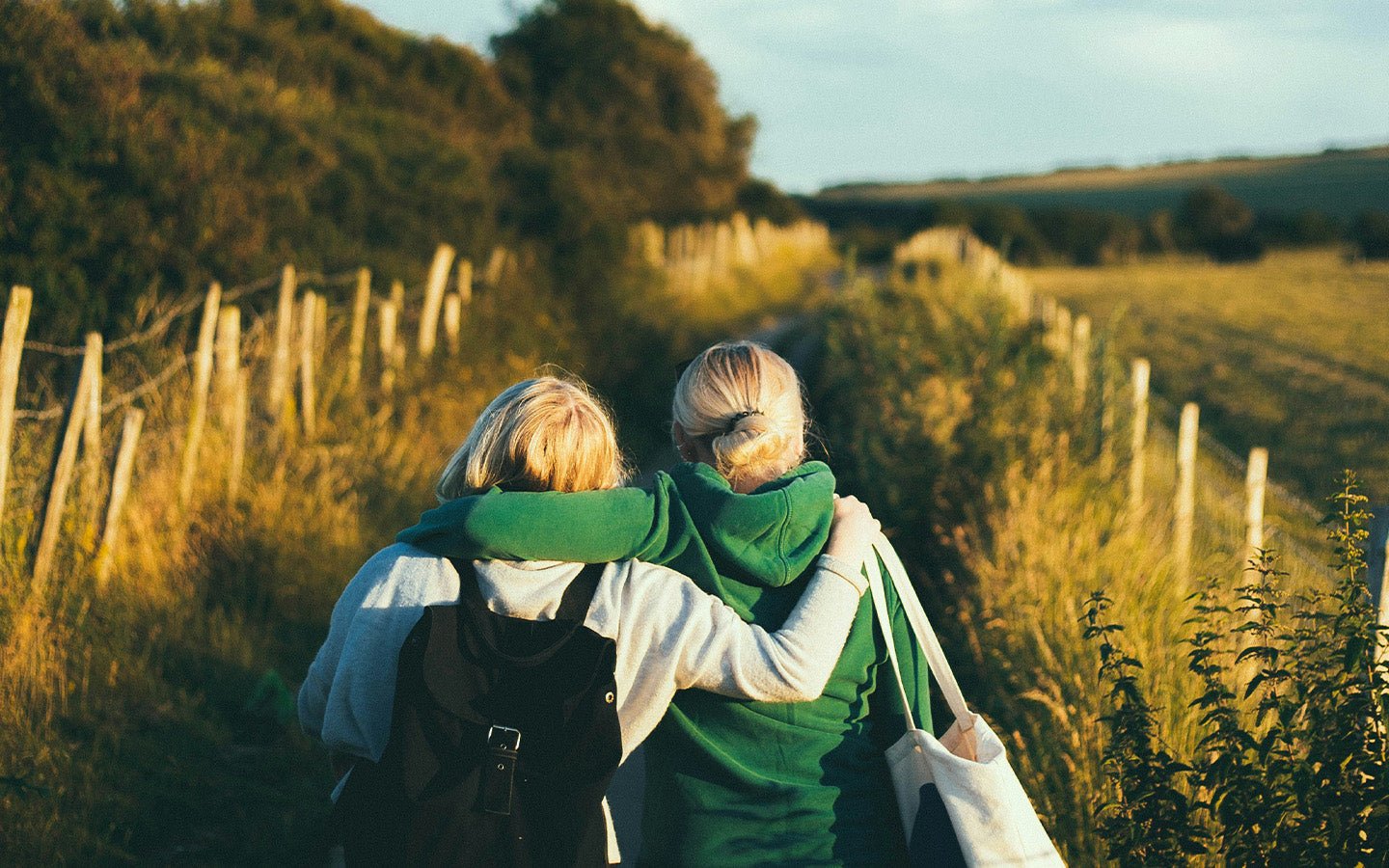 Two women in green hoodies walk arm-in-arm on a path next to a fence and fields