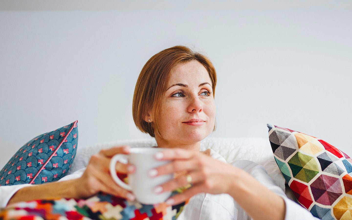 Woman with short brown hair holding a white mug, sitting on a white couch with colorful pillows