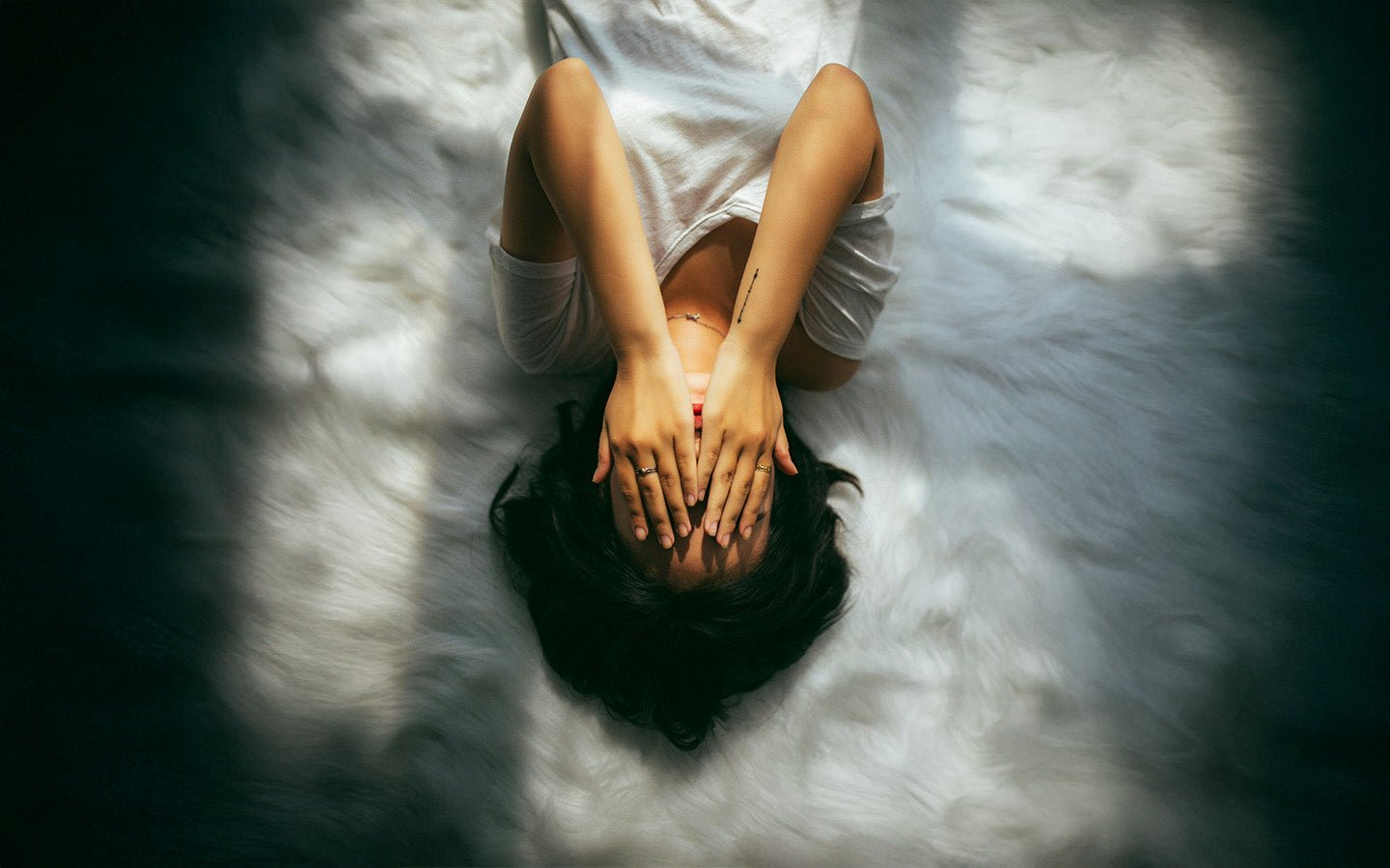 Woman lying on white faux fur rug, hands covering face, sunlight and shadow