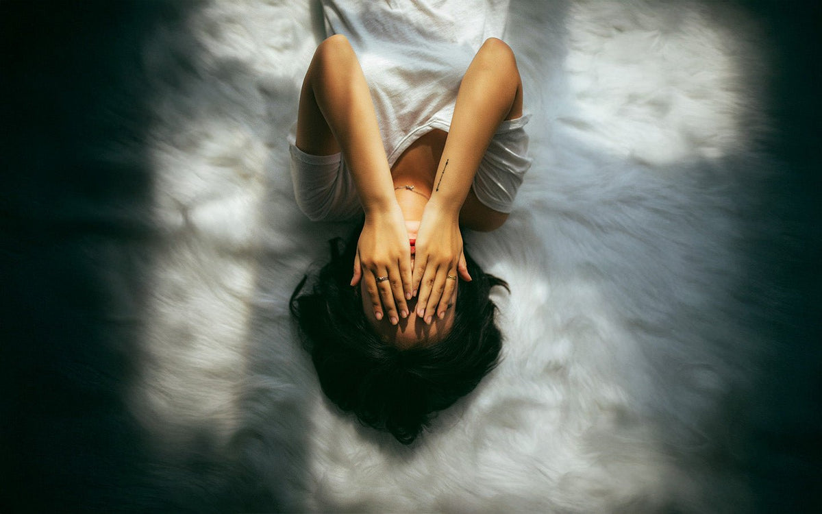 Woman lying on white faux fur rug, hands covering face, sunlight and shadow