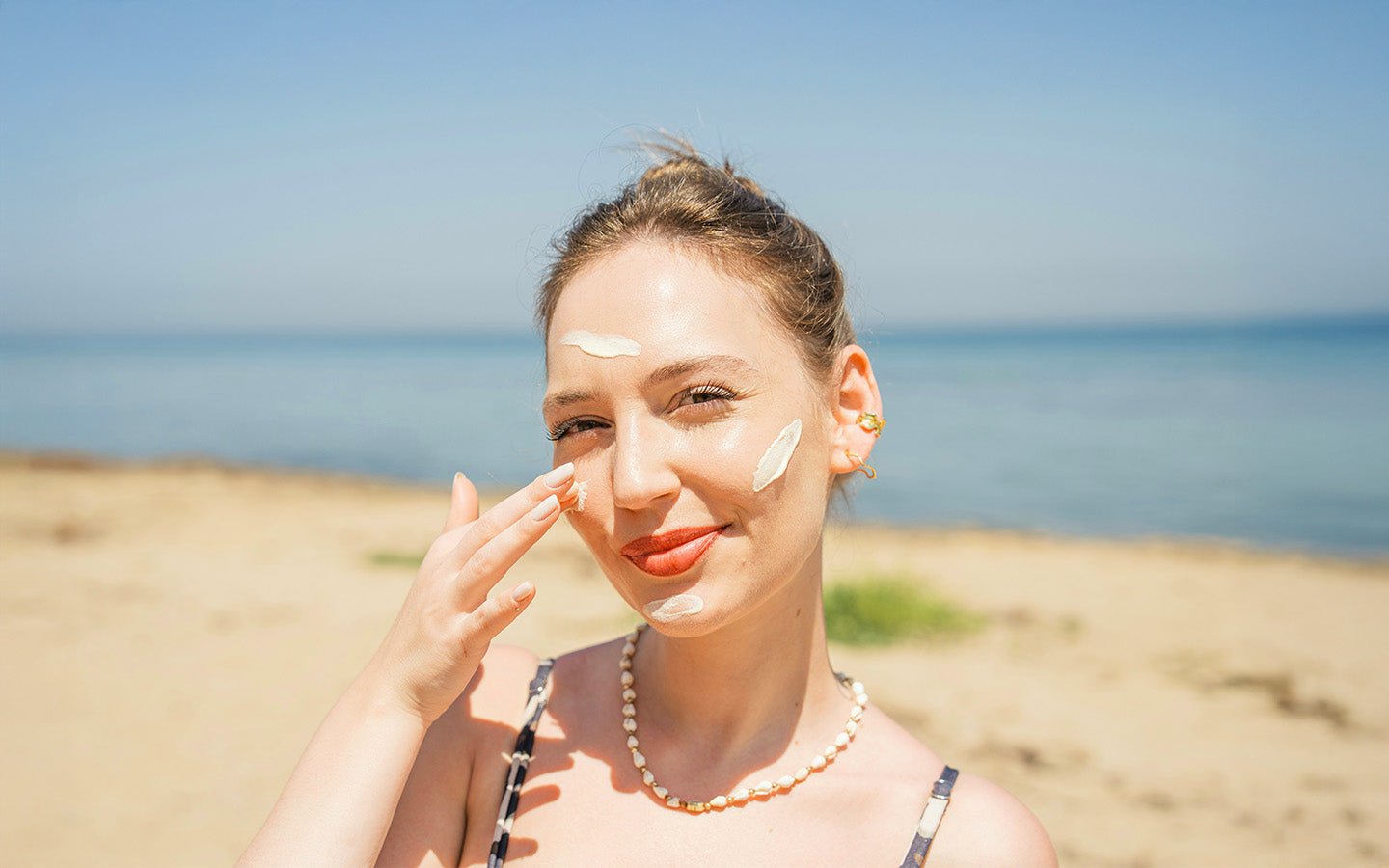Woman on beach applying white sunscreen to her face, wearing a pearl necklace