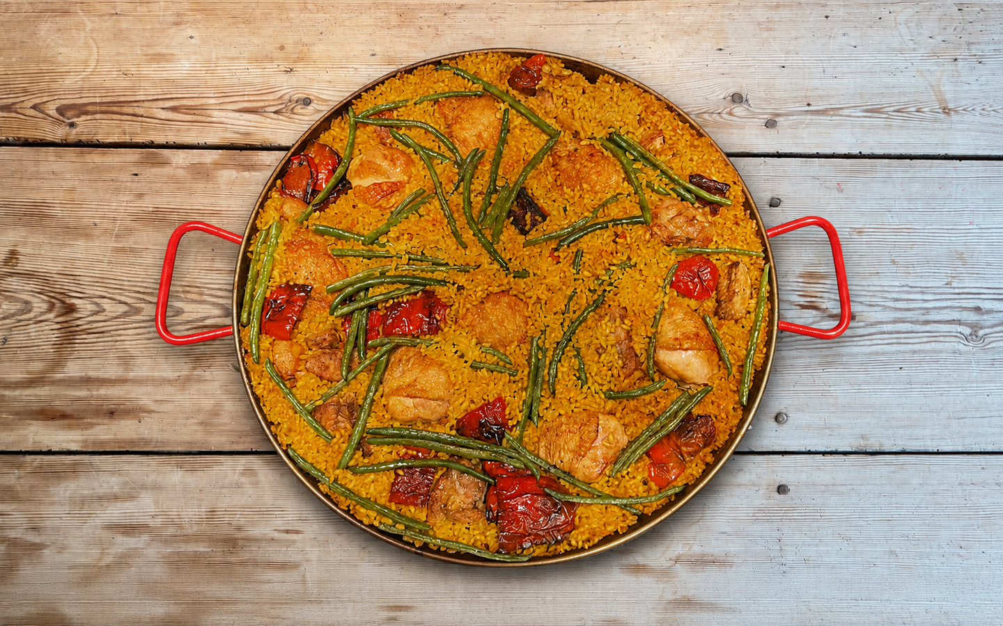 Overhead view of paella with chicken, green beans and red peppers in a red-handled pan on a wooden kitchen table.
