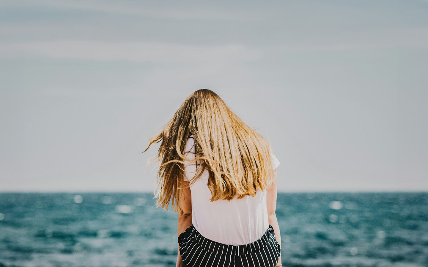 Woman with long blonde hair, white shirt, and striped pants facing the ocean
