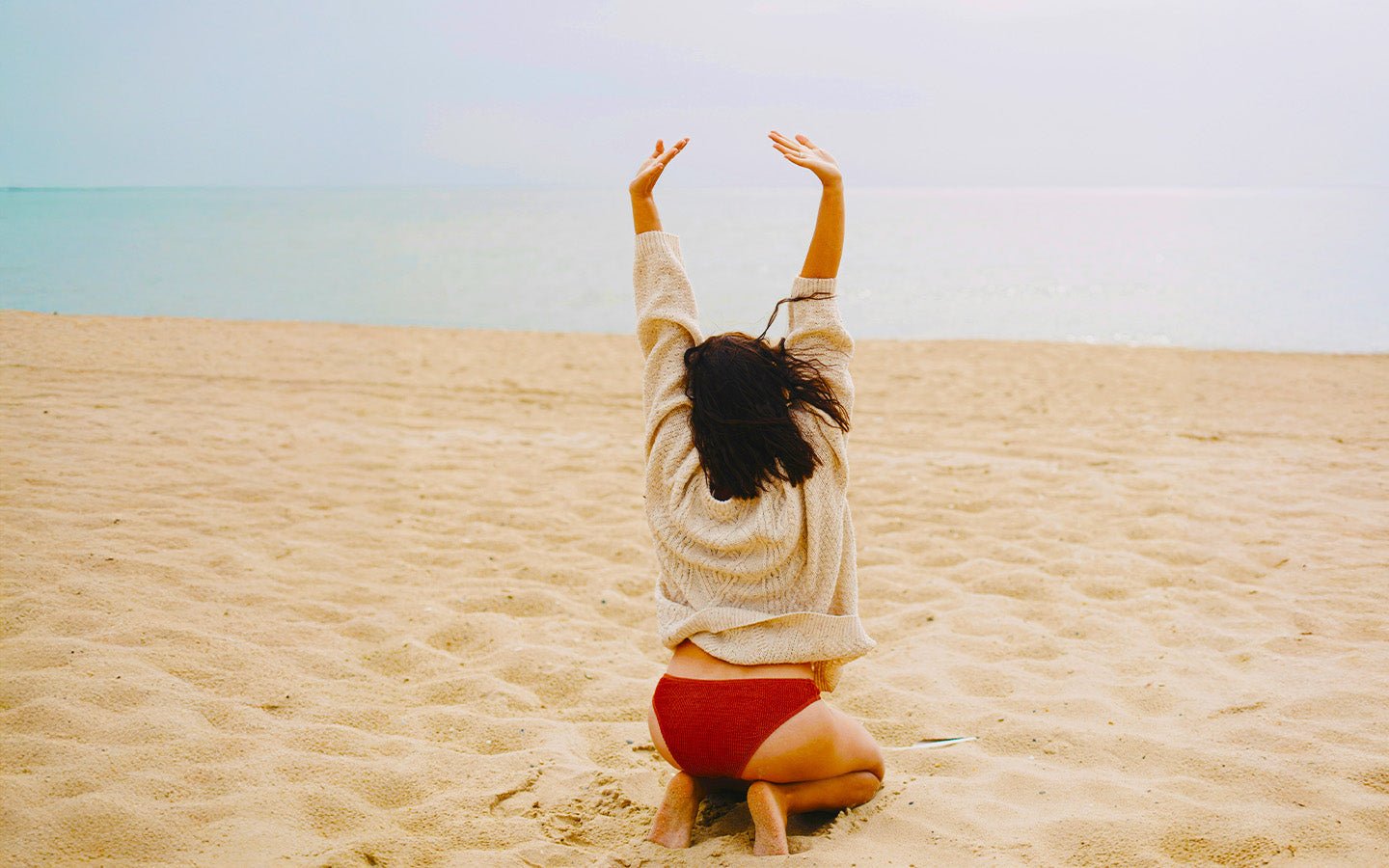 Woman in beige sweater and red bikini bottoms stretching on a sandy beach