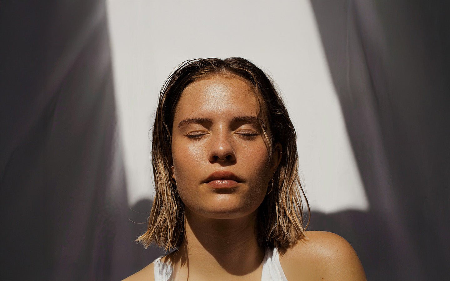 Young woman with closed eyes and wet hair, sunlit against a white and gray wall