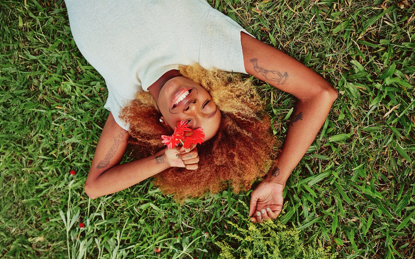 Woman with red hair and tattoos smiles, holding red flowers, lying on green grass