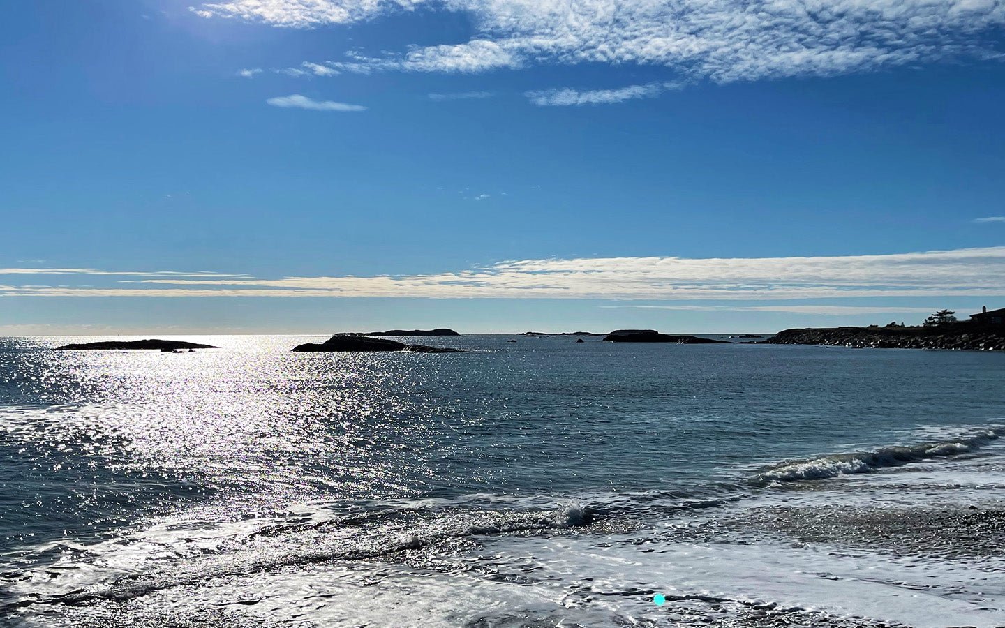 Sunlight sparkling on calm ocean water with a rocky shoreline under a blue sky