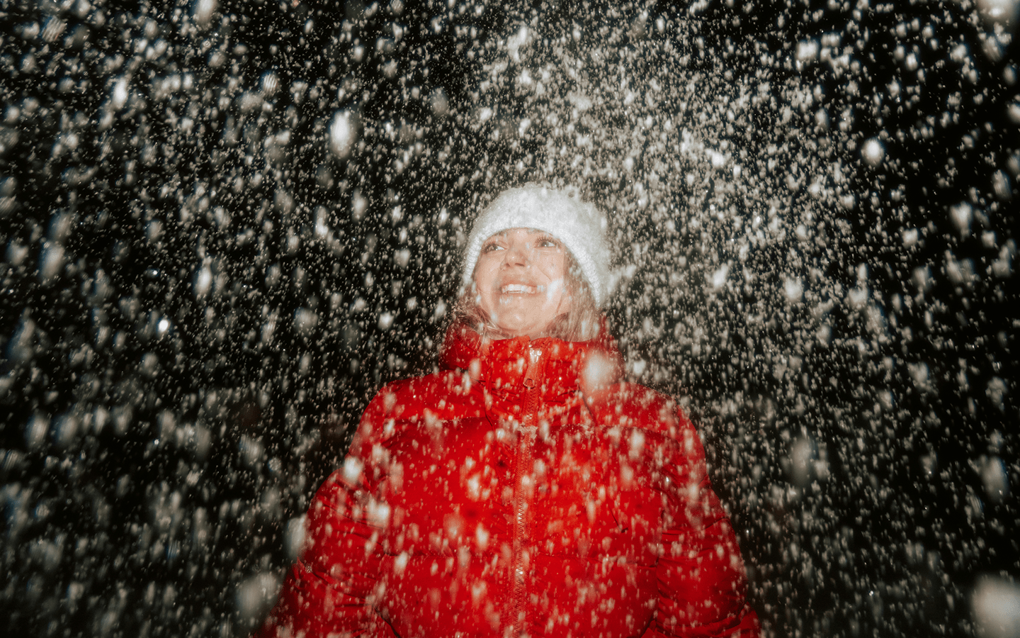Woman in red coat and white hat smiling up at falling snow