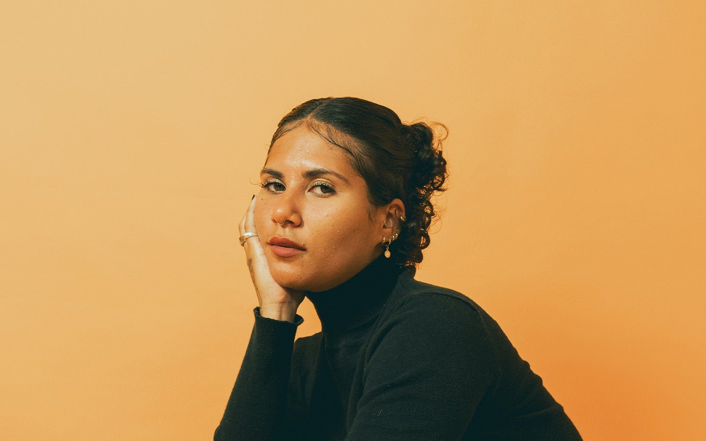 Woman with dark hair in a bun, black turtleneck, and nose ring, resting chin on hand